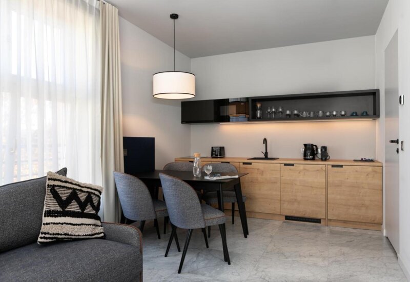Aneks kuchenny w apartamencie. Meble są drewniane a przed nimi stoi czarny stół na cztery osoby z szarymi krzesłami. Z przodu po lewej widać fragment szarej kanapy z poduszką.