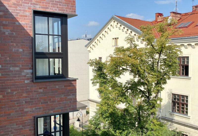 Widok z okna. Po lewej róg pokrytego cegłą budynku, z prawej kremowy budynek z czerwoną dachówką.