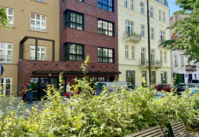 Na pierwszym planie drewniane ławki otoczone przez zielone krzewy. W tle kamienice, z wyróżniającym się, pokrytym cegłą budynkiem pośrodku.