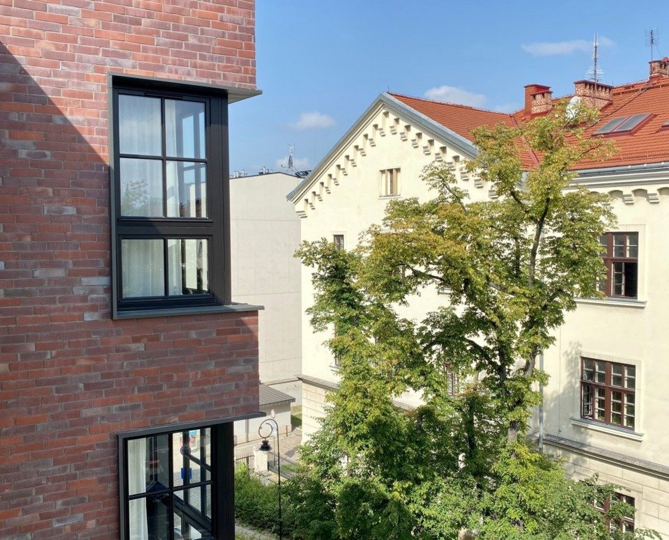 Widok z okna. Po lewej róg pokrytego cegłą budynku, z prawej kremowy budynek z czerwoną dachówką.