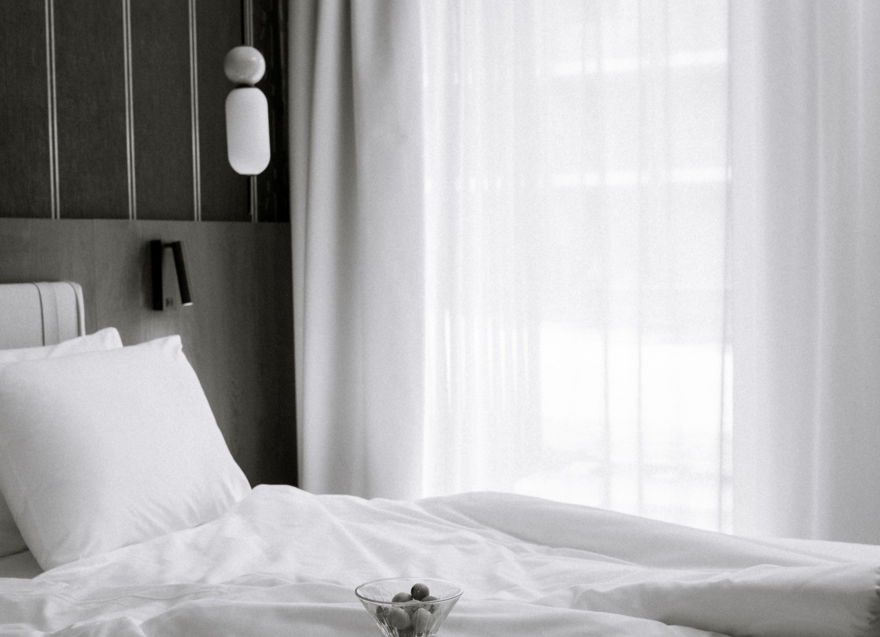 Jasny, nowoczesny pokój hotelowy z pogniecionym białym łóżkiem, lekkimi zasłonami, lampą i tacą z kieliszkiem, przekąską i czasopismami.