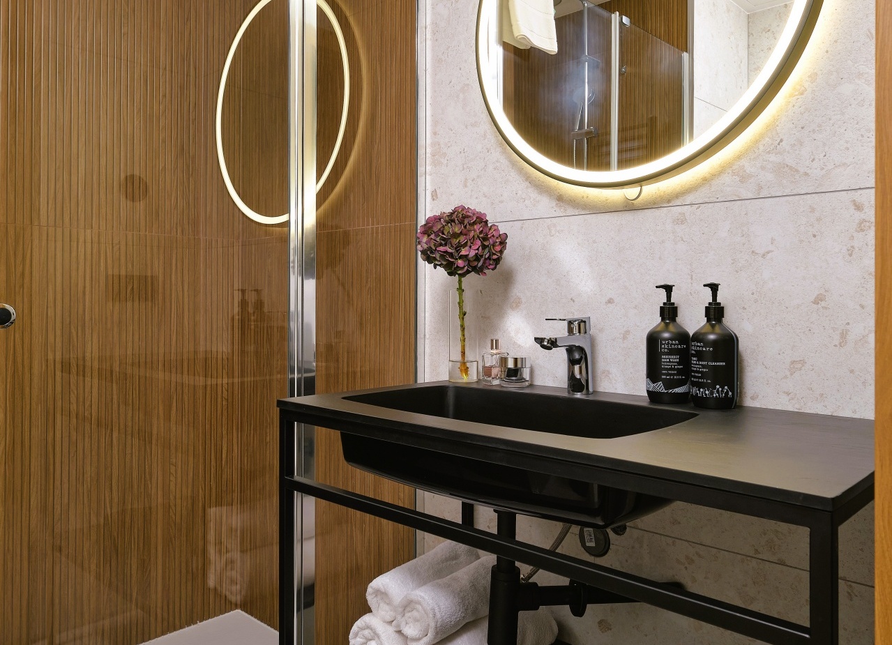 Nowoczesna łazienka hotelowa z prysznicem, drewnianą ścianą, podświetlanym okrągłym lustrem, czarną umywalką i zwiniętymi ręcznikami.