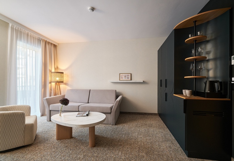 Příjemný moderní salon v hotelovém apartmá s béžovou pohovkou a křeslem, mramorovým stolkem, stojací lampou, závěsy a minibarem s konvicí.