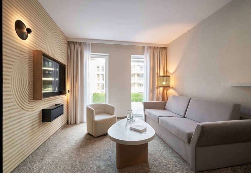 Světlé lounge apartmá s neutrální pohovkou, křeslem, mramorovým stolkem s vodou, nástěnnou TV a okny od podlahy ke stropu