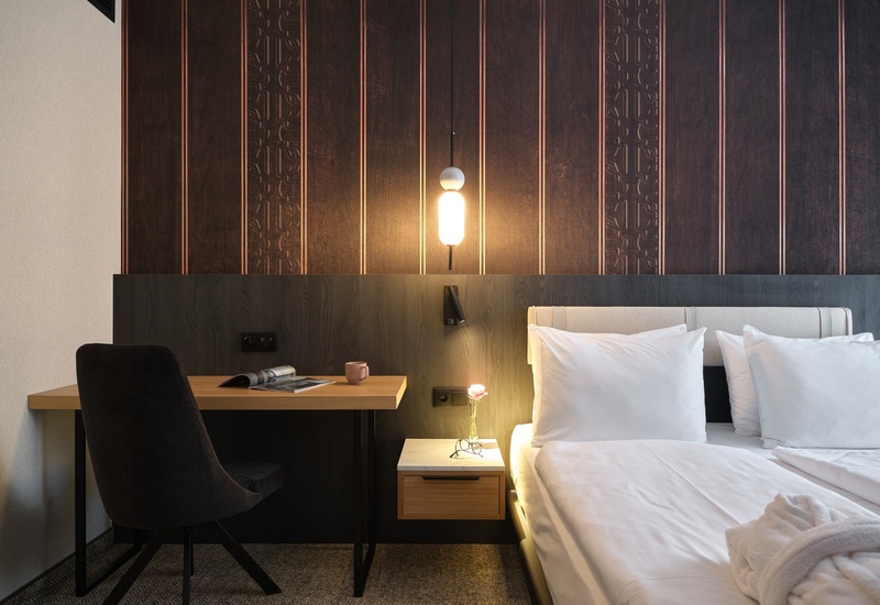 Moderní hotelový pokoj s dvojlůžkem a bílým povlečením, teplým závěsným světlem, tmavým dřevěným čelem a plovoucím nočním stolkem s růží