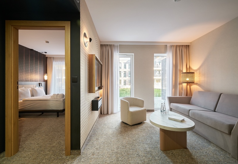 Moderní hotelový apartmán s obývací částí: pohovka, křeslo, mramorový konferenční stolek, televize na stěně, teplé světlo, velká okna a přilehlá ložnice.