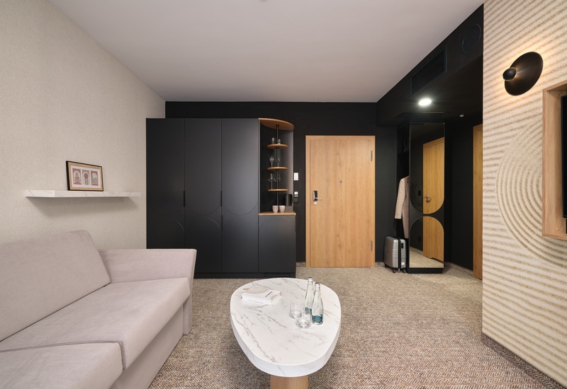 Moderní hotelový koutek s béžovou pohovkou, mramorovým stolkem s lahvemi vody, matně černou skříní, dřevěnými dveřmi a zavazadlem