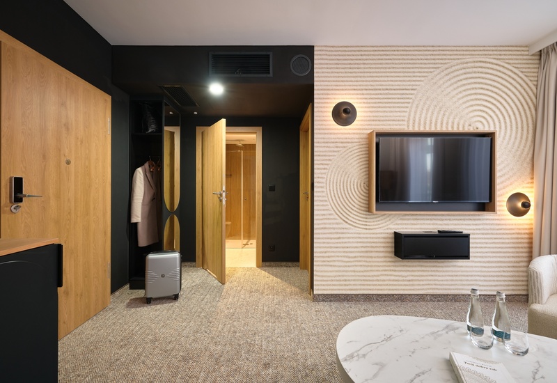 Moderní vstup do pokoje s dřevěnými dveřmi, otevřenou skříní a zavazadlem, strukturovanou dekorativní stěnou, TV a mramorovým stolkem s vodou.