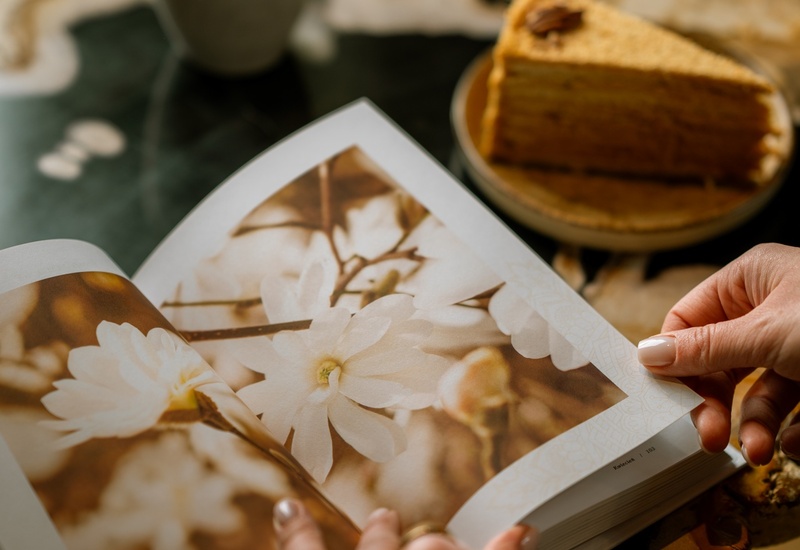 Dłonie przeglądające album ze zdjęciami kwiatów na stoliku, obok filiżanka herbaty i kawałek ciasta