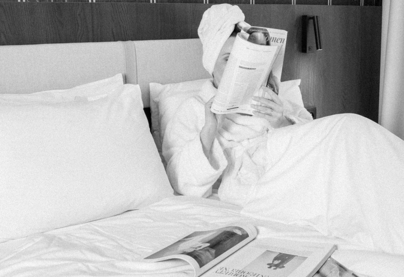 Host v županu s ručníkem na hlavě čte časopis na bílém hotelovém lůžku s polštáři, dřevěným čelem a noční lampičkou.