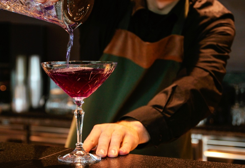 Barman w ciemnej koszuli i fartuchu nalewa intensywnie fioletowy koktajl z kryształowego shakera do eleganckiego kieliszka coupe w barze hotelowym