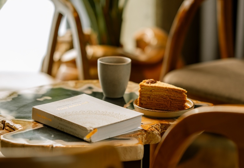 Przytulny kącik hotelowy: drewniany stolik i krzesła, filiżanka kawy, kawałek ciasta i książka przy doniczkowej roślinie