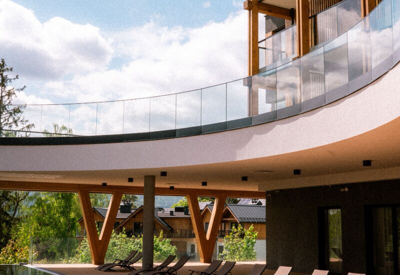 Zadaszony basen spa z drewnianymi belkami, zakrzywioną szklaną balustradą i rzędami leżaków przy lustrującej tafli wody pod błękitnym niebem.