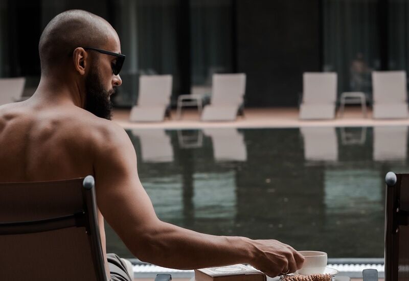 Man in sunglasses relaxing on a lounger at the hotel pool; cup, book and cookies on a side table, empty loungers reflected in water