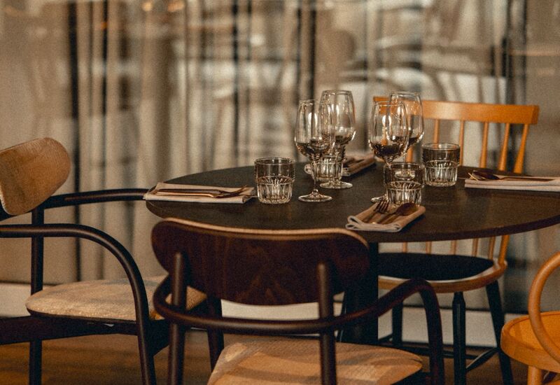Stół w restauracji zastawiony kieliszkami do wina, szklankami i sztućcami; drewniane krzesła i miękkie światło przez lekkie zasłony.