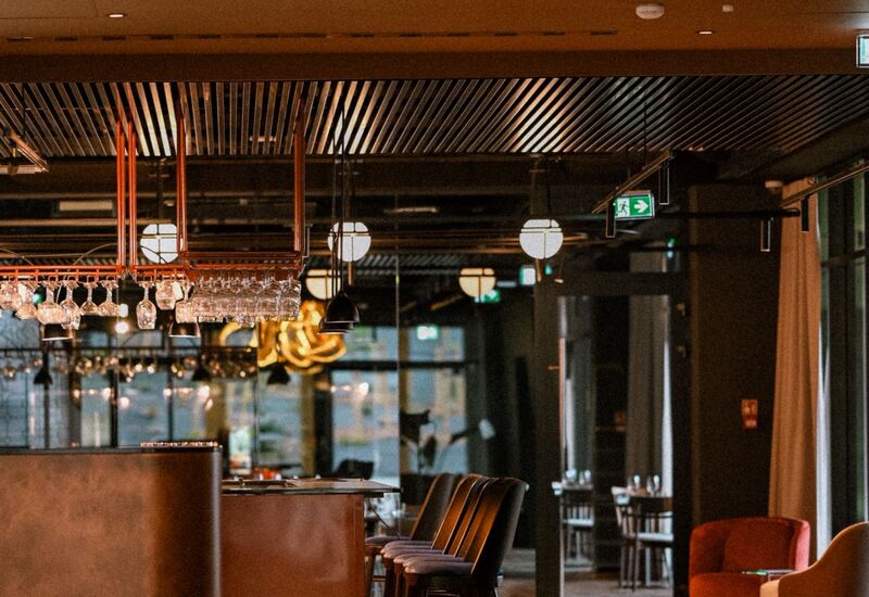 Przytulny bar hotelowy z wysokimi hokerami, wiszącymi kieliszkami, ciepłym oświetleniem i miejscami do relaksu.
