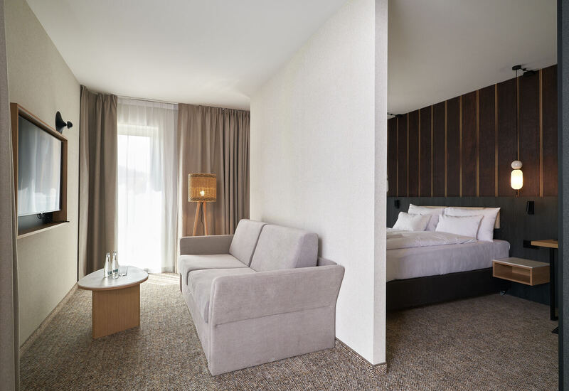 Nowoczesny apartament z salonem: szara sofa, stolik z wodą, pleciona lampa stojąca; sypialnia z białą pościelą i ciemną drewnianą ścianką.