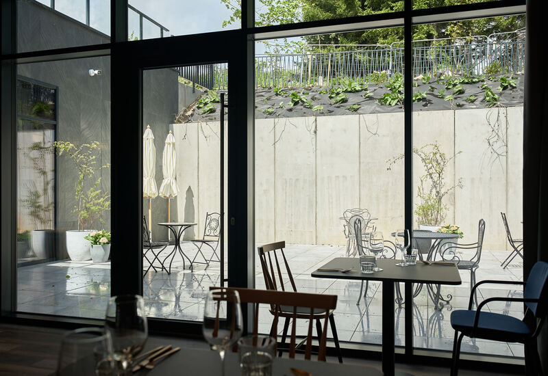 Jadalnia wewnątrz przy przeszklonej ścianie z widokiem na słoneczne patio ze stolikami bistro, parasolami i donicami