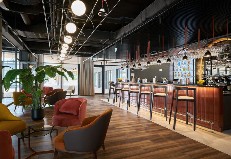 Stylowy hotelowy bar i lounge z wysokimi stołkami, wiszącymi lampami, półkami na kieliszki, przytulnymi fotelami i drewnianą podłogą.