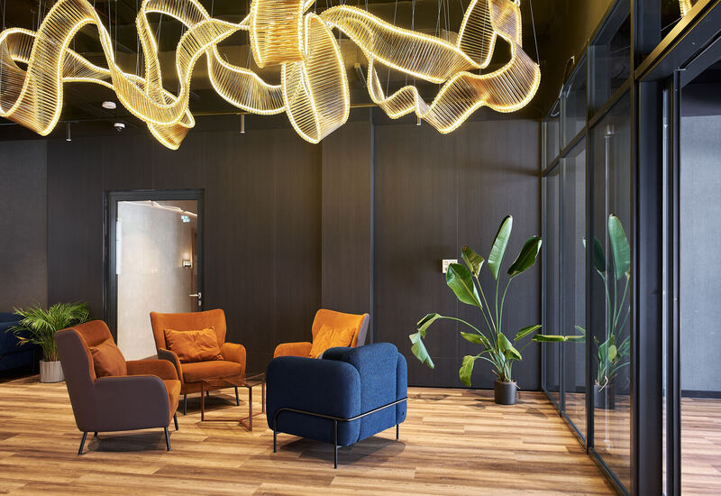 Nowoczesny salon hotelowy z rzeźbiarskim złotym żyrandolem, pomarańczowymi i granatowymi fotelami, drewnianą podłogą i roślinami.