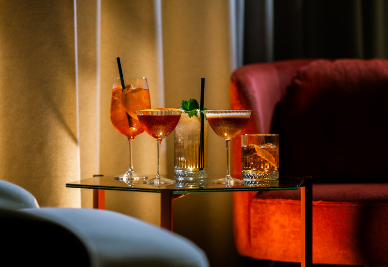 Koktajle na szklanym stoliku w przytulnym hotelowym lounge, różne szkła, ozdoby cytrusowe i mięta, czerwony welurowy fotel w tle