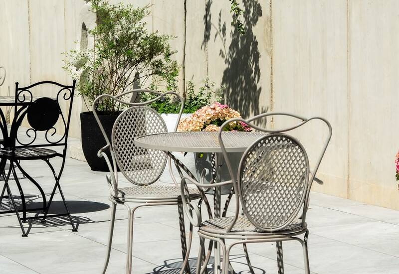 Słoneczne patio z ozdobnym metalowym stolikiem bistro i krzesłami, donice z krzewami i kwiatami przy jasnej ścianie