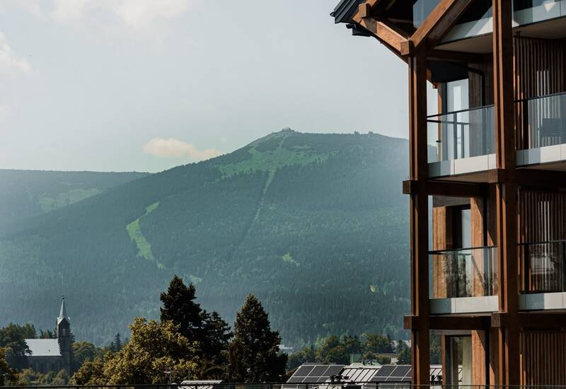 Chalet-style hotel with timber beams and glass balconies overlooking forested mountain slopes and a distant church spire