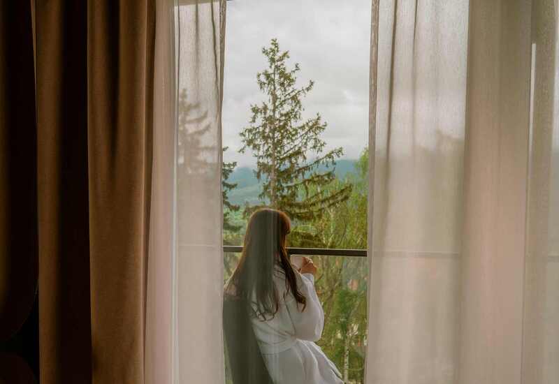 Guest in a white robe on a balcony holding coffee, framed by sheer curtains with pine trees and mountain view