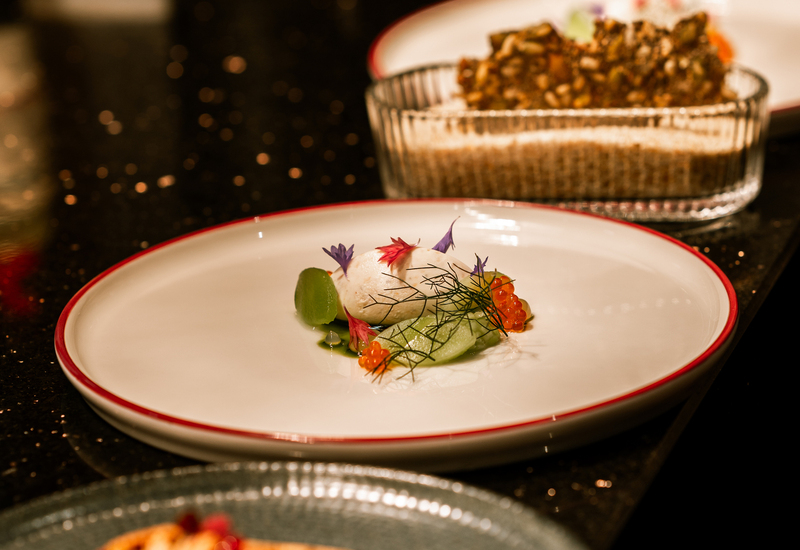 Amuse-bouche szefa: kulka z owoców morza z ikrą łososia, plasterkami ogórka, koperkiem i jadalnymi kwiatami na białym talerzu; chrupiący dodatek z tyłu.