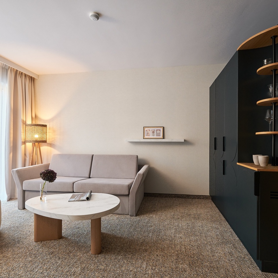 Przytulny, nowoczesny salon w apartamencie hotelowym z beżową sofą, fotelem, marmurową ławą, lampą i minibarem z czajnikiem.