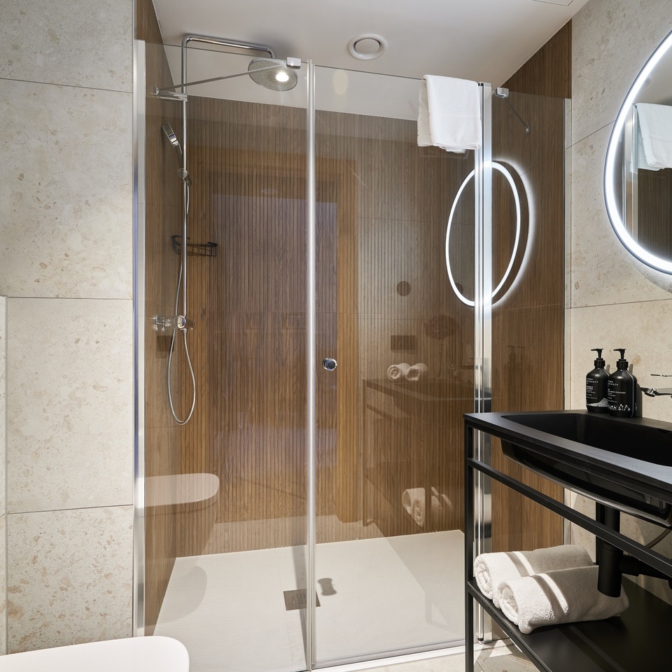 Nowoczesna łazienka hotelowa z przeszklonym prysznicem walk-in, deszczownicą i słuchawką, drewnianą ścianą, podświetlanym lustrem i ręcznikami