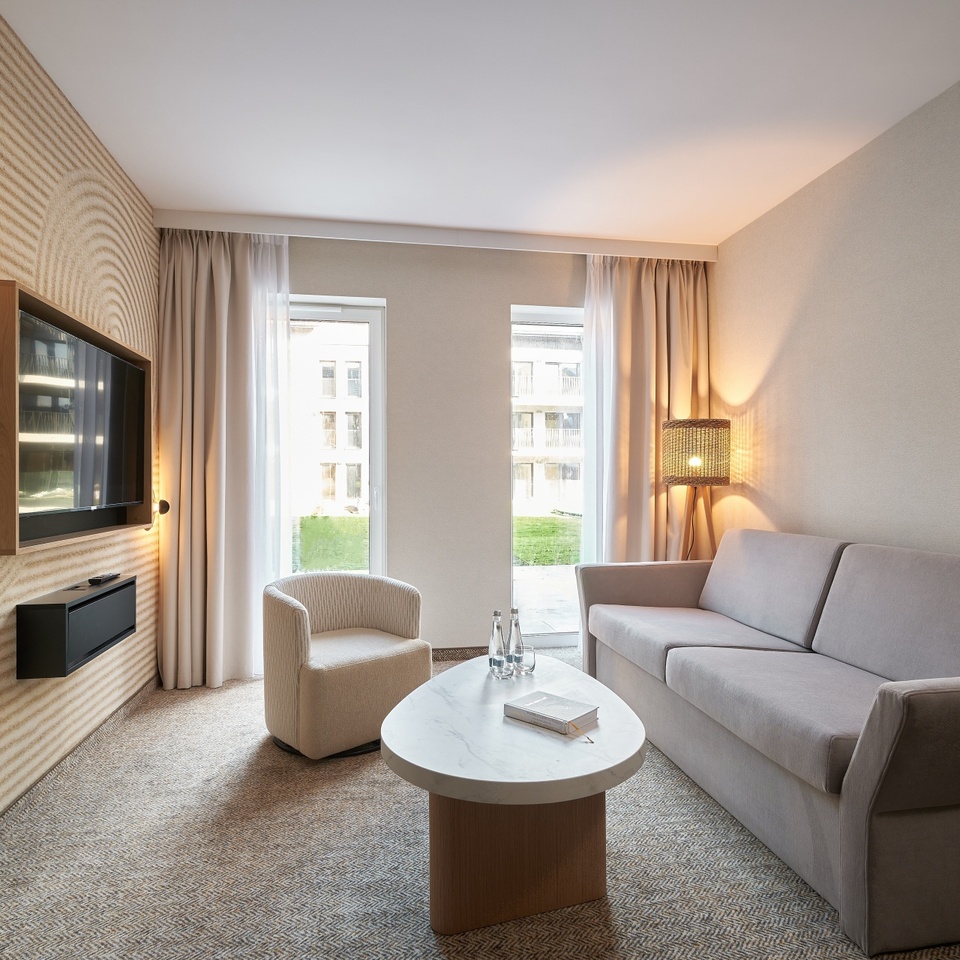 Jasny salon apartamentu z neutralną sofą, fotelem, marmurową ławą z wodą, TV na ścianie i oknami od podłogi do sufitu