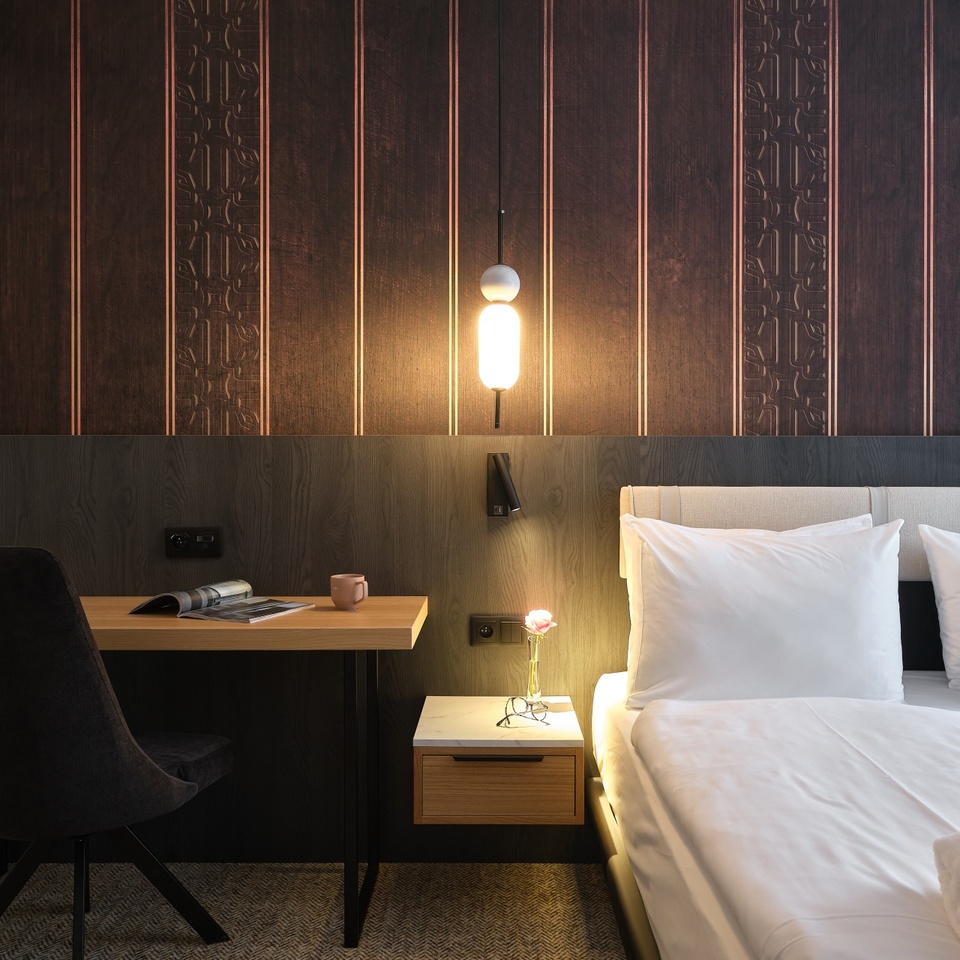 Nowoczesny pokój hotelowy z podwójnym łóżkiem i białą pościelą, ciepłą lampą wiszącą, ciemnym drewnianym zagłówkiem i stolikiem z różą