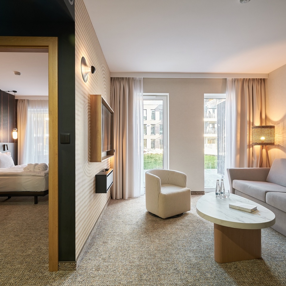 Nowoczesny apartament z salonem: sofa, fotel, marmurowy stolik, telewizor na ścianie, ciepła lampa stojąca, duże okna i przyległa sypialnia.