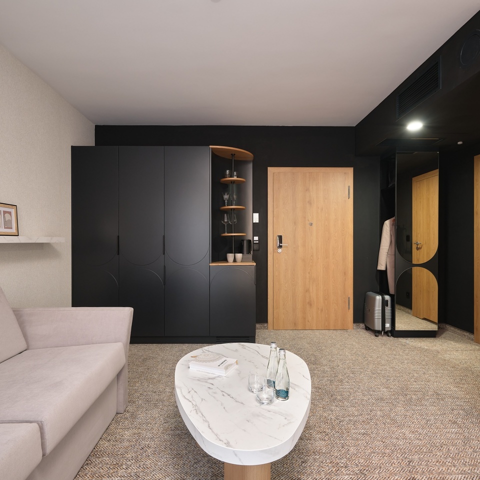 Nowoczesny kącik hotelowy z beżową sofą, marmurowym stolikiem z butelkami wody, matową czarną szafą, drewnianymi drzwiami i walizką
