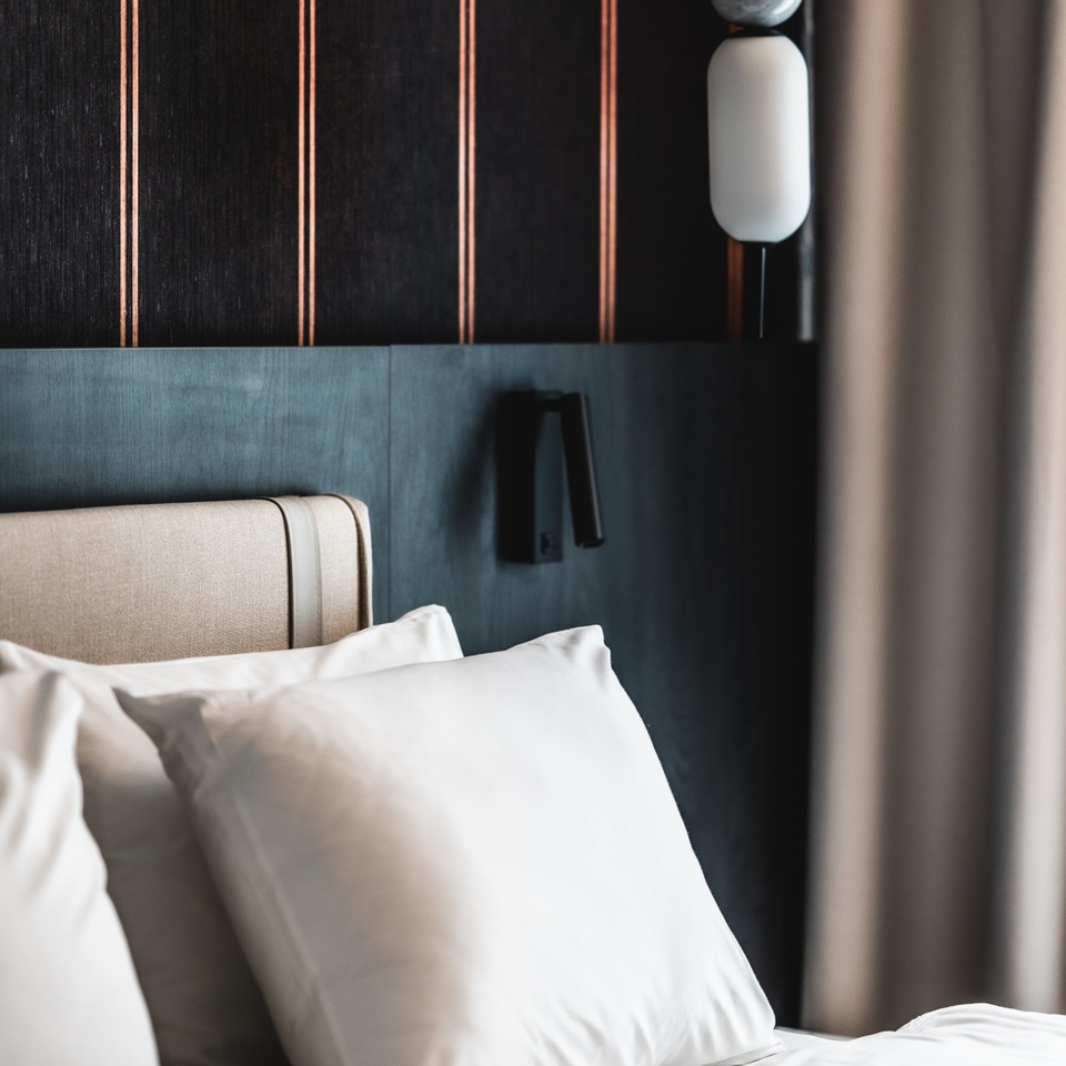 Miękkie białe poduszki i świeża pościel na nowoczesnym łóżku z beżowym zagłówkiem, ciemną, paskowaną ścianą i lampą wiszącą