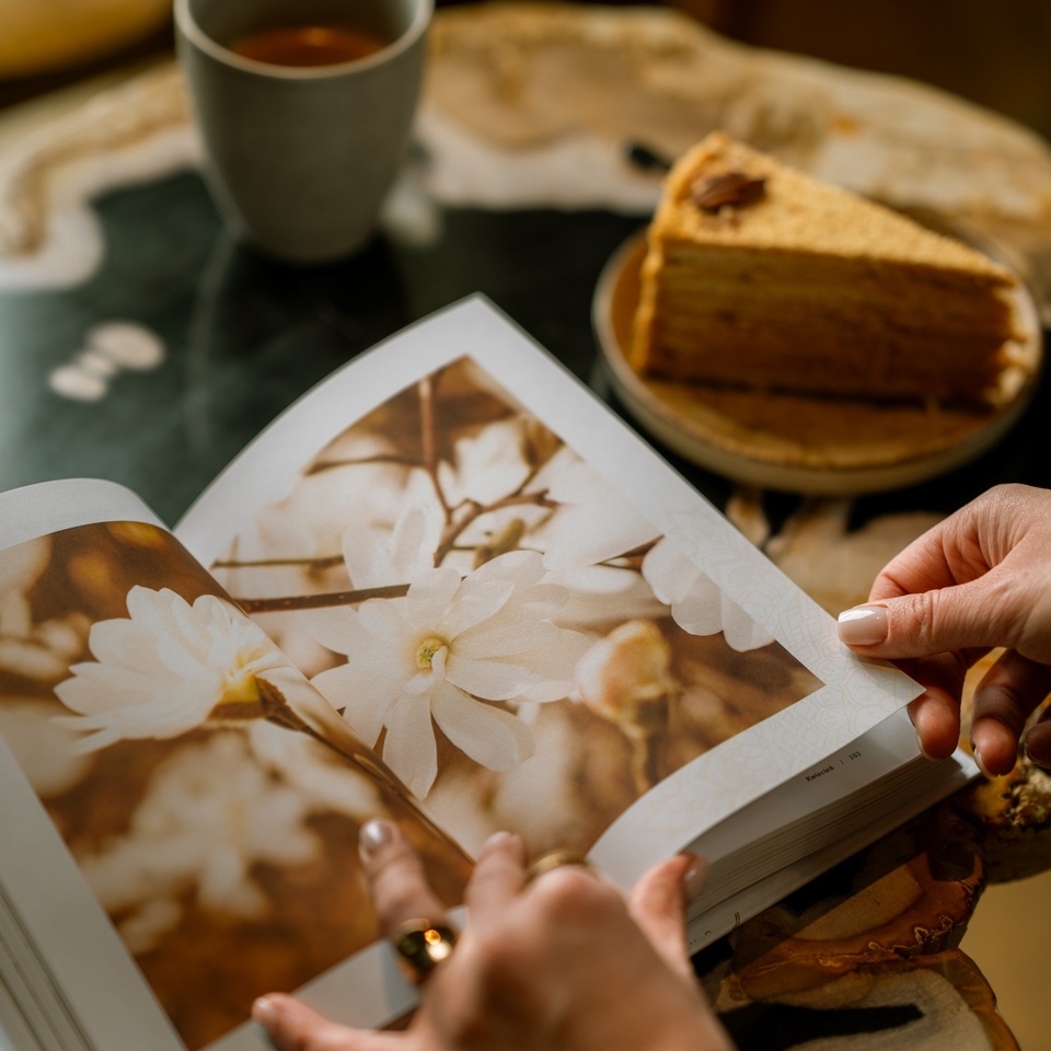 Dłonie przeglądające album ze zdjęciami kwiatów na stoliku, obok filiżanka herbaty i kawałek ciasta