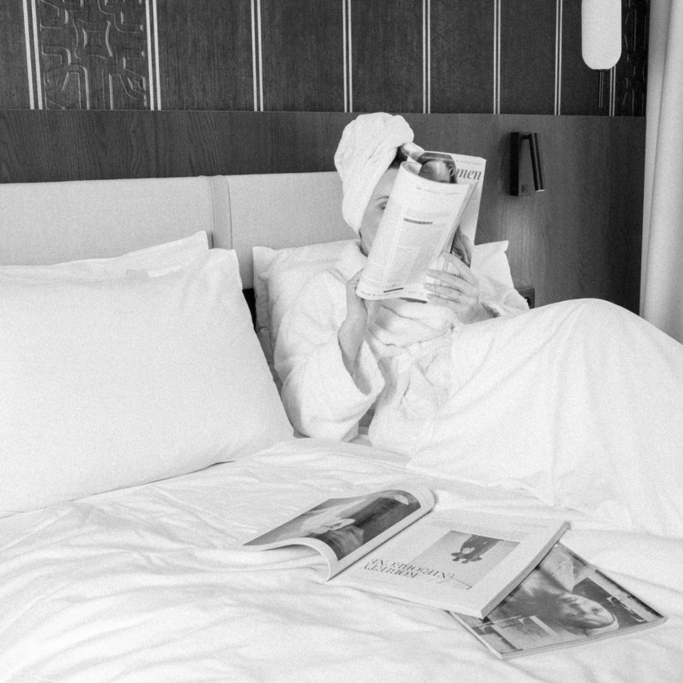 Gość w szlafroku i ręczniku na głowie czyta magazyn na białym hotelowym łóżku z poduszkami, drewnianym zagłówkiem i lampką nocną.