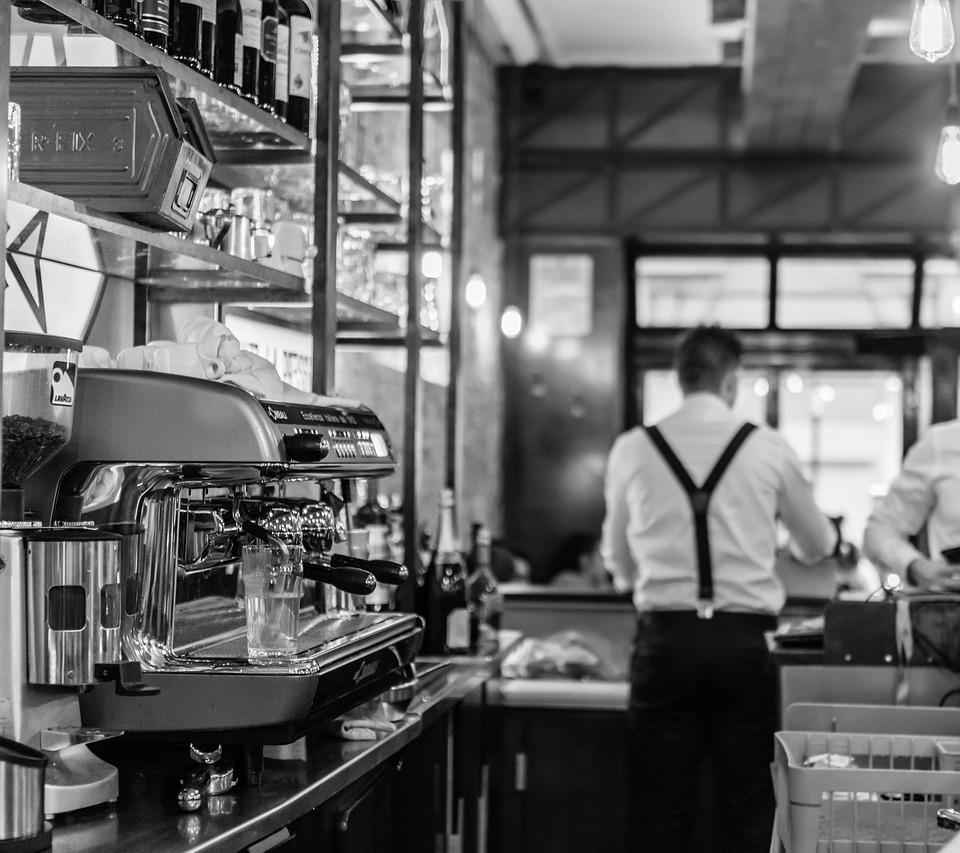 Polerowany chromowany ekspres do kawy na barze z przeszklonymi półkami, personel w szelkach przygotowuje napoje