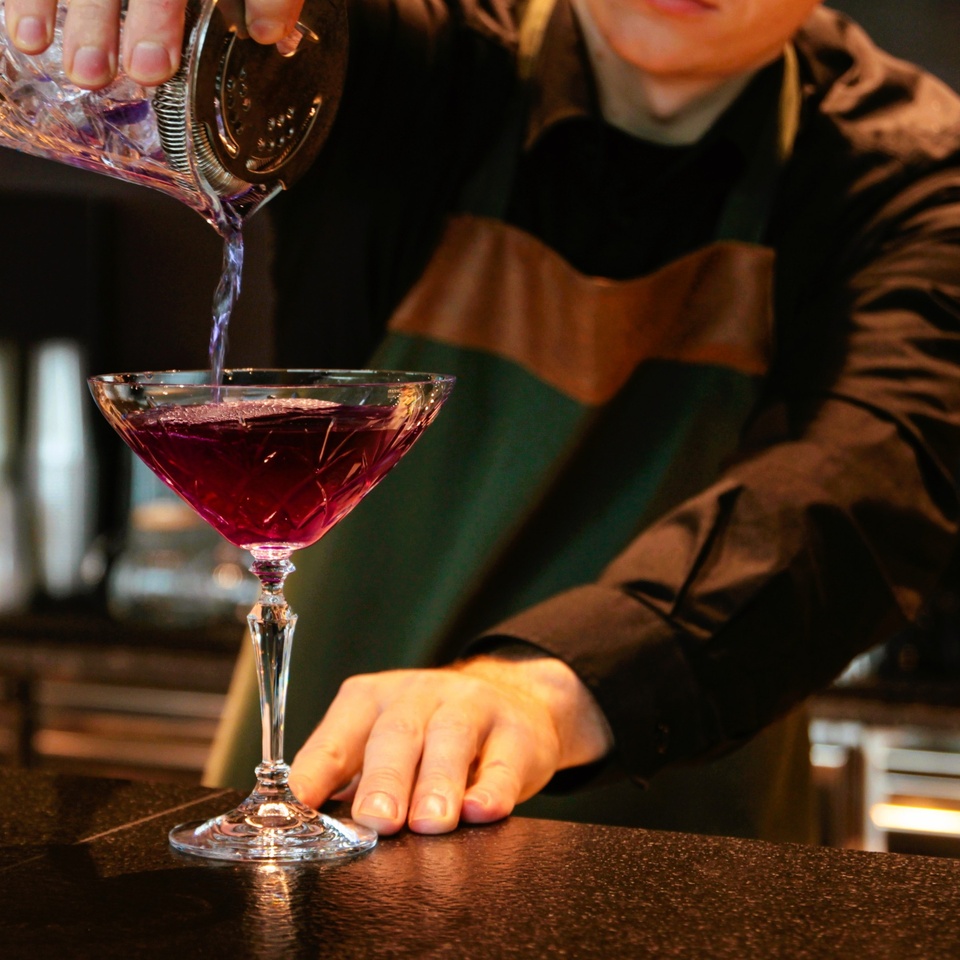 Barman w ciemnej koszuli i fartuchu nalewa intensywnie fioletowy koktajl z kryształowego shakera do eleganckiego kieliszka coupe w barze hotelowym