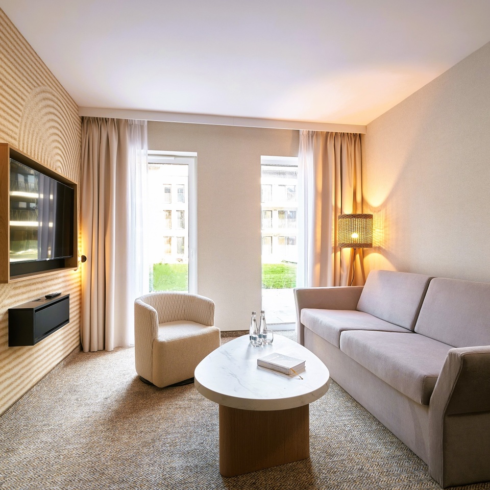 Jasny hotelowy salon z sofą i fotelem, marmurowym stolikiem z butelkami wody i książką, telewizorem i widokiem na dziedziniec