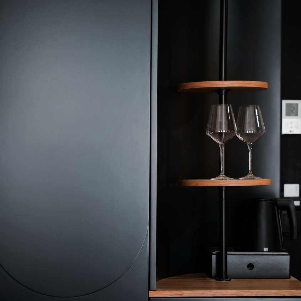 Matowa, czarna wnęka minibaru z drewnianymi półkami, dwoma kieliszkami i czajnikiem — nowoczesne udogodnienie hotelowe.