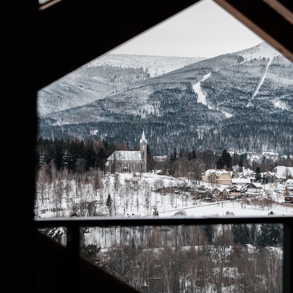 Widok z balkonu obramowany ciemnymi drewnianymi belkami na zaśnieżoną wieś z wieżą kościoła, odbicie na jeziorze i stoki narciarskie