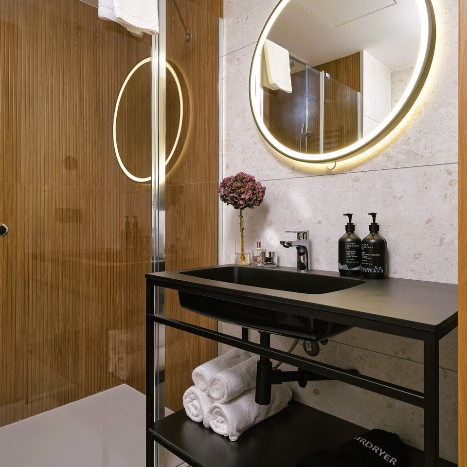 Nowoczesna łazienka hotelowa z prysznicem, drewnianą ścianą, podświetlanym okrągłym lustrem, czarną umywalką i zwiniętymi ręcznikami.