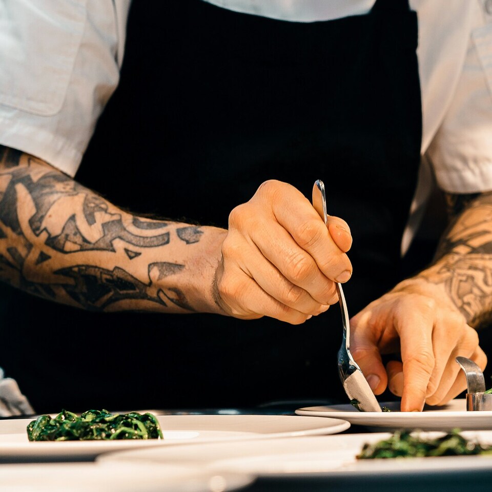Kucharz z wytatuowanymi ramionami w białej koszuli i czarnym fartuchu precyzyjnie nakłada duszoną zieleninę na białe talerze