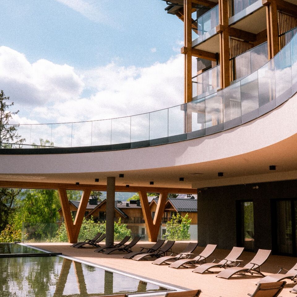 Zadaszony basen spa z drewnianymi belkami, zakrzywioną szklaną balustradą i rzędami leżaków przy lustrującej tafli wody pod błękitnym niebem.