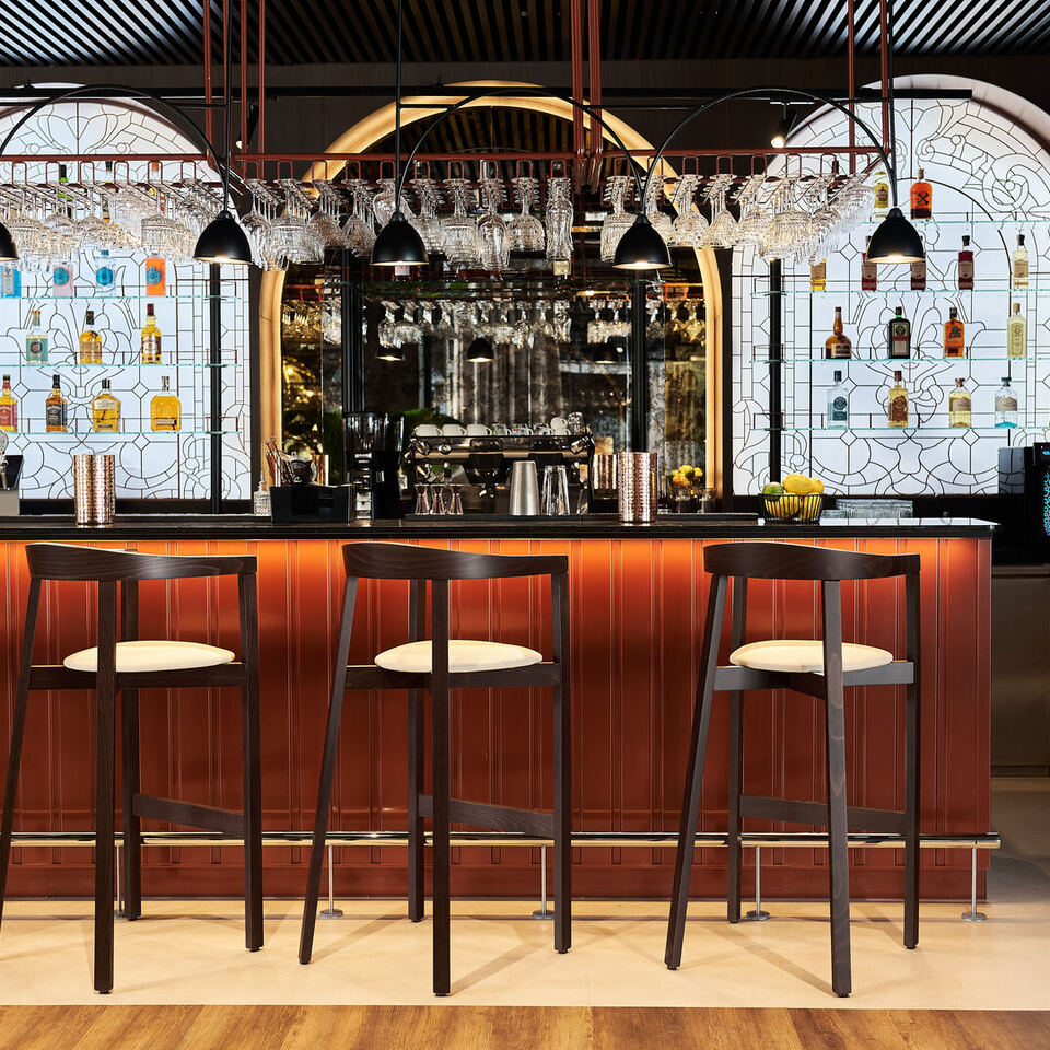 Stylowy bar hotelowy z czterema drewnianymi stołkami, miedzianym blatem, wiszącymi kieliszkami i podświetlonymi półkami z butelkami.