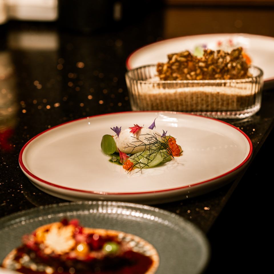 Amuse‑bouche šéfkuchaře: kulička z mořských plodů s lososovým kaviárem, plátky okurky, koprem a jedlými květy na bílém talíři; semínkový křupánek vzadu.