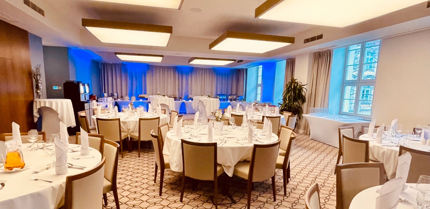 Elegant hotel banquet room with round tables, white linens, folded napkins, buffet station, soft ceiling lights and blue uplighting.