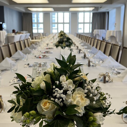 Long banquet table with white linens, folded napkins, glassware and elegant white floral centerpieces in a bright event room